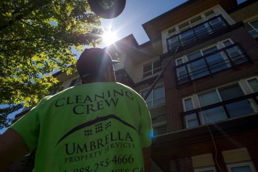 A man with a shirt that says 'cleaning crew Umbrella' on the back. He is holding a long washing brush on a pole against the windows of a low-rise building